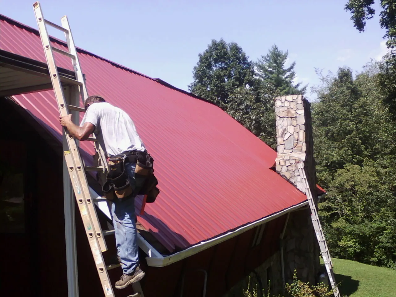 Roofer on a ladder beside a red metal roof and stone chimney — Soffit & Fascia Repair by Roofing Brothers in Garnett